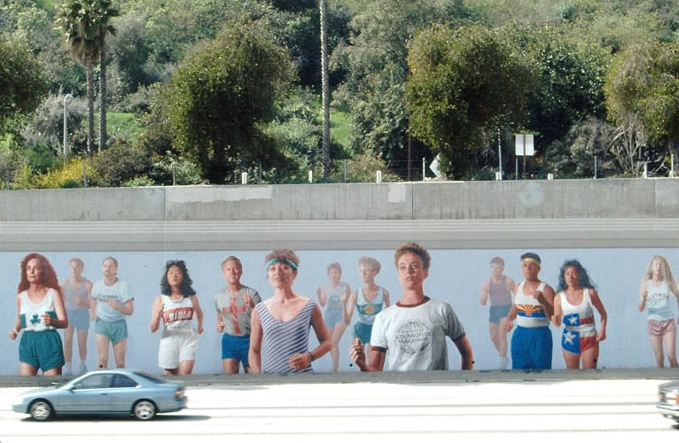 Close-up of the LA Marathon Mural, showing several life-size painted runners on a freeway retaining wall with palm trees and hills behind